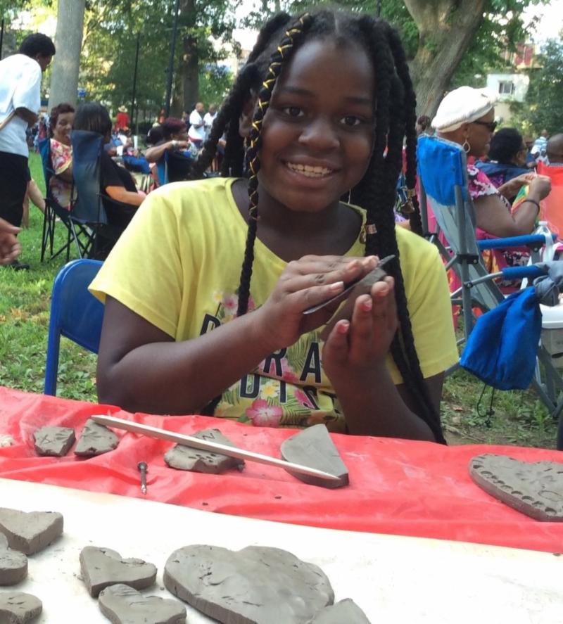 A young girl with braided hair smiles while shaping clay at a community event, surrounded by other people sitting in the background.