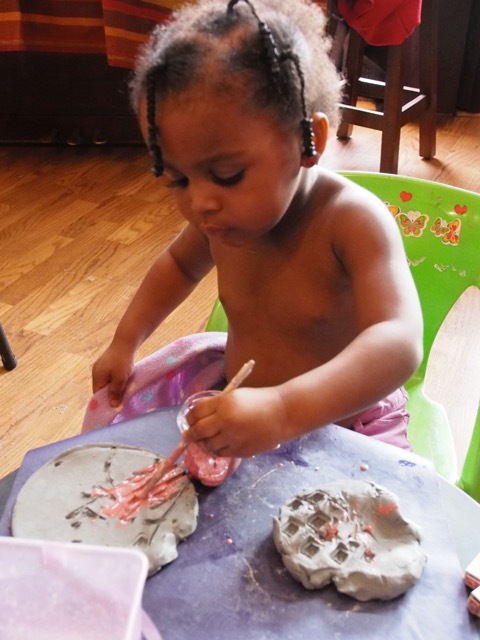 A young child with curly hair is creatively playing with clay on a table, using a paintbrush to apply pink color to the gray clay shapes.