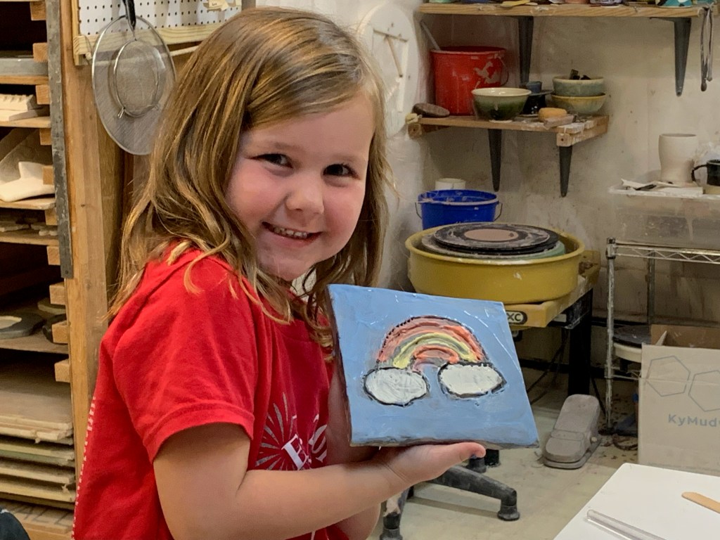 A smiling young girl is holding a painted tile featuring a rainbow and clouds in a pottery studio.