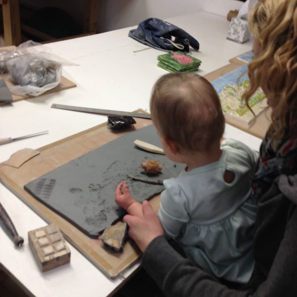 A child sitting on the lap of an adult, both engaged in a crafting activity at a table. Various art supplies, including clay and tools, are scattered around them.