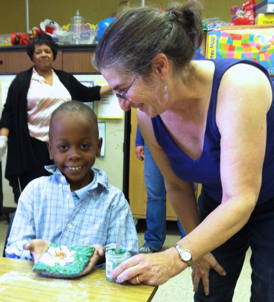 A smiling child holds a painted art piece on a table while an adult woman leans in closely, smiling at the child. Another woman is visible in the background.