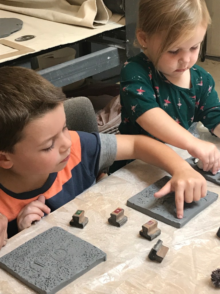 Two children engaged in a creative activity, placing colorful stamps onto gray clay tiles on a table. The boy is focused on his work, while the girl is carefully adjusting her stamp.