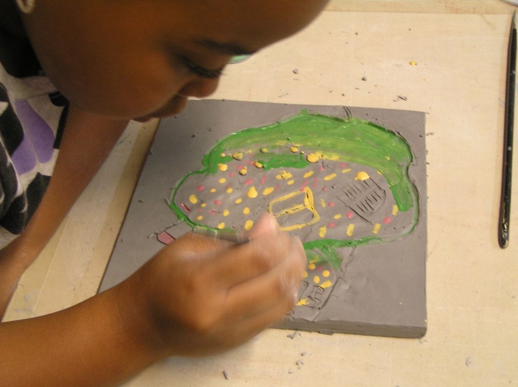 A child painting a colorful design on a gray clay slab, featuring green, yellow, and pink details.