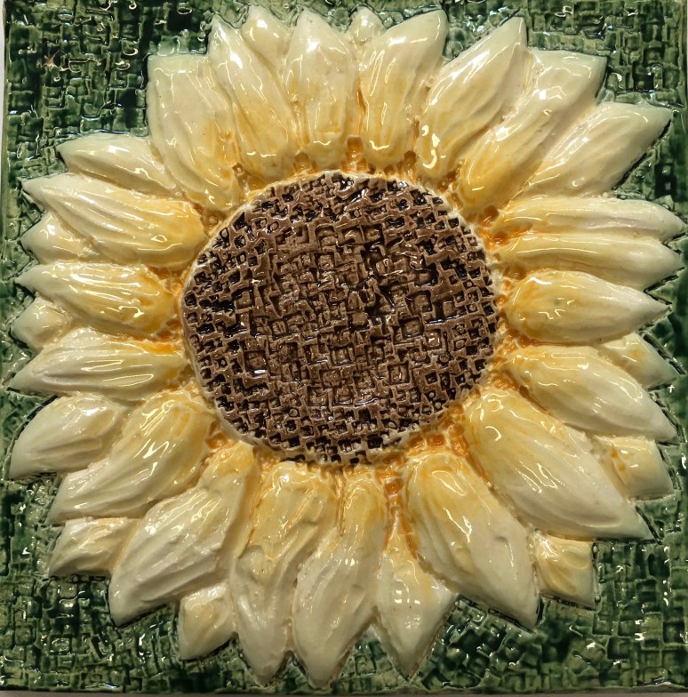 A decorative ceramic tile featuring a sunflower with a textured brown center and light yellow petals, surrounded by a green background.