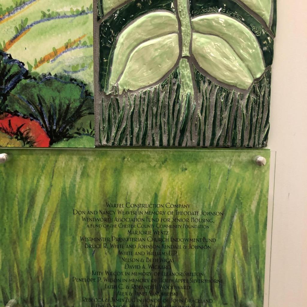 A decorative plaque featuring a green and brown relief design of leaves and grass, alongside inscriptions of names and memorials in white text.